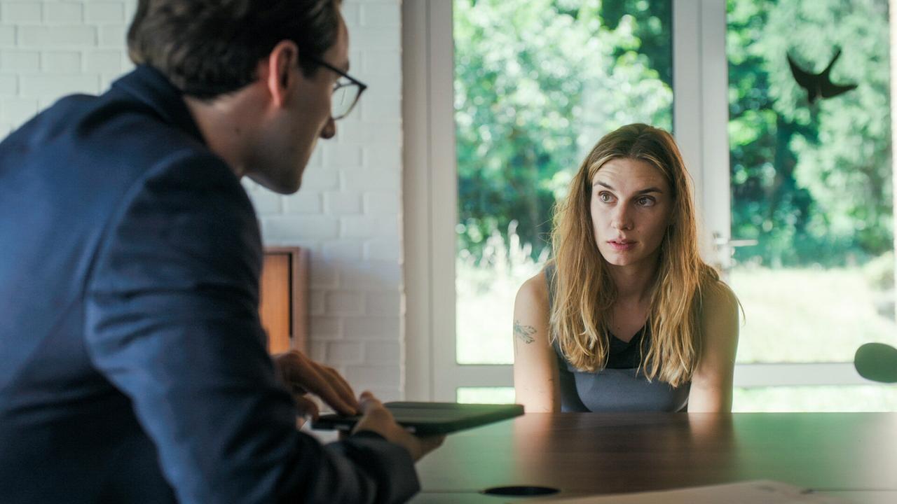 Tobias Resch befragt Maya Unger an einem dunklem Holztisch,  im Hintergrund große Fenster mit Blick ins Grüne.