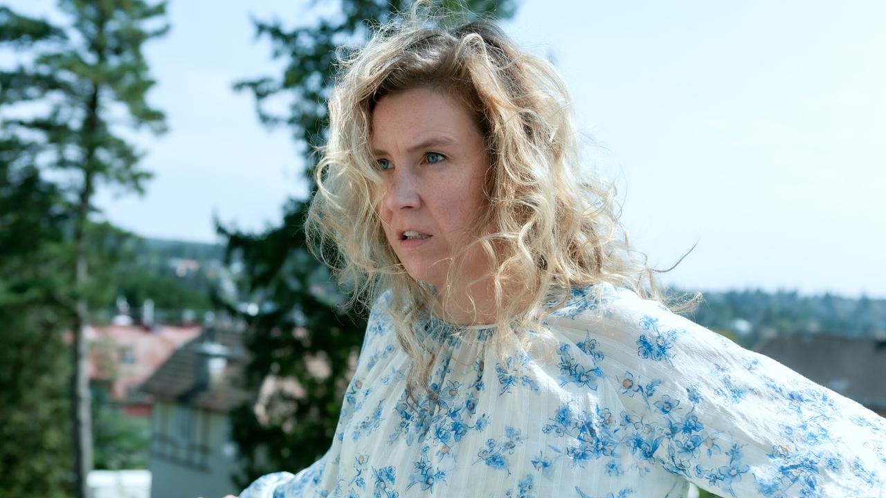 Franziska Hackl in weißer Bluse mit mit blauem Blumenmuster steht auf einer Dachterrasse, im Hintergrund sind Bäume und Dächer zu sehen.