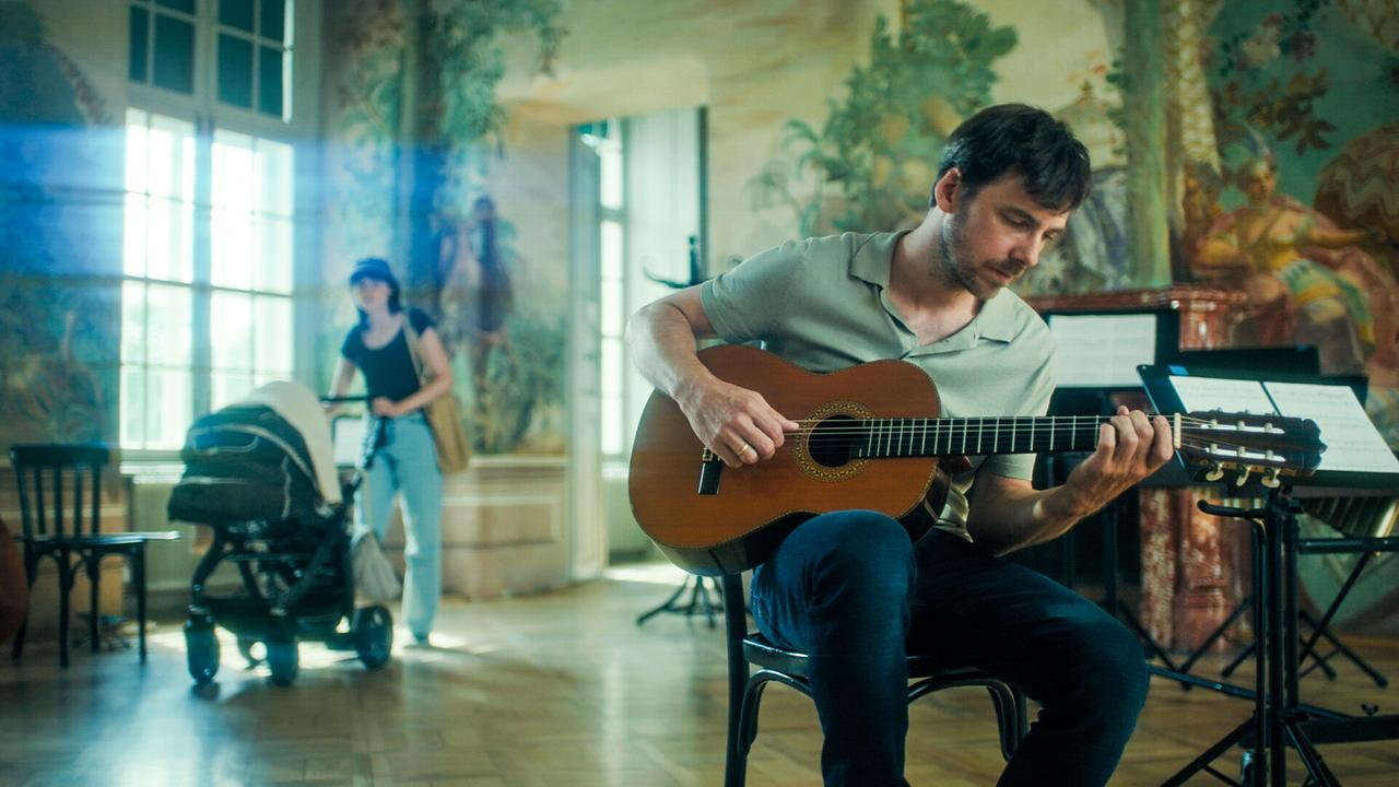 Stefan Pohl sitzt auf einem Sessel einem in Raum mit Wandmalereien und spielt Gitarre, während im Hintergrund Paulina Hobratschk mit einem Kinderwagen steht.