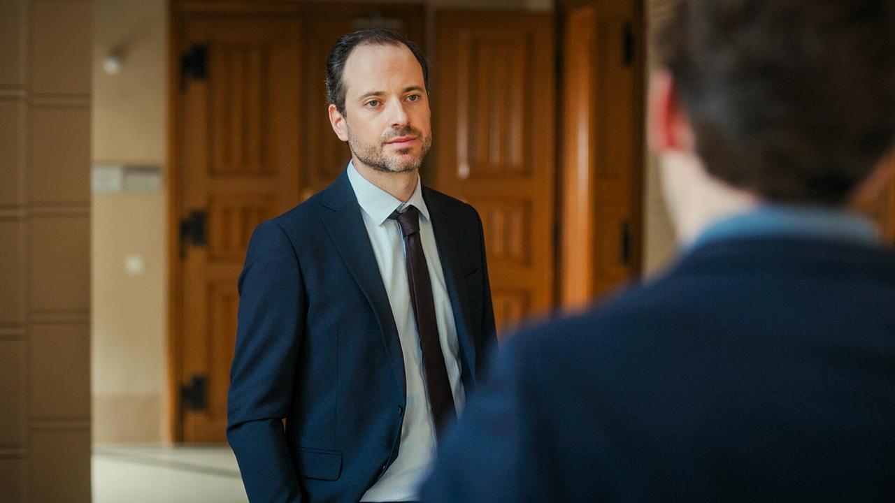 Alexander Absenger im Anzug steht vor einer weiteren Person in einem Gebäude mit großen Holztüren.