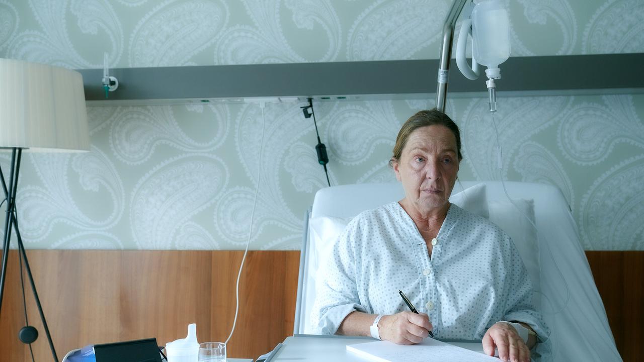 Susi Stach sitzt aufrecht in einem Krankenhausbett und schreibt auf einem Blatt Papier, neben ihr medizinische Ausrüstung.