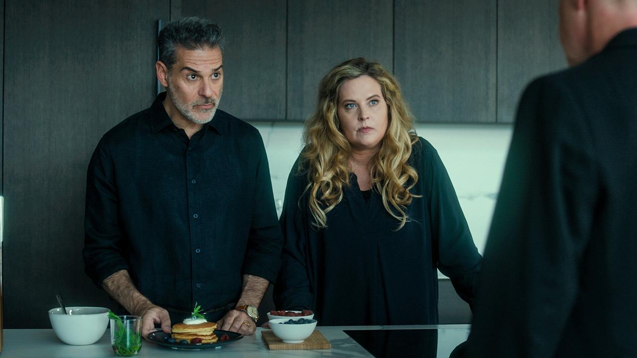 Rick Kavanian und Diana Amft blicken in einer modernen Küche mit vorbereiteten Speisen und Getränken auf der Arbeitsfläche auf eine weitere Person ihnen gegenüber.