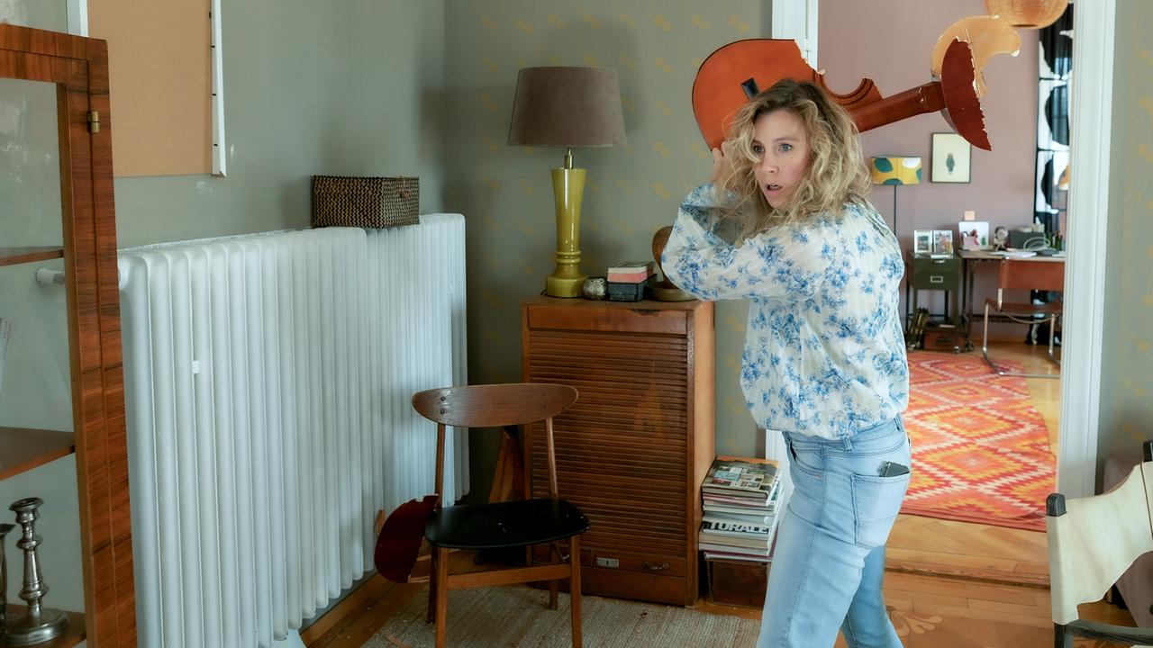 Franziska Hackl hält in einem Wohnzimmer eine zerbrochene Gitarre über den Kopf, als wolle sie diese werfen.