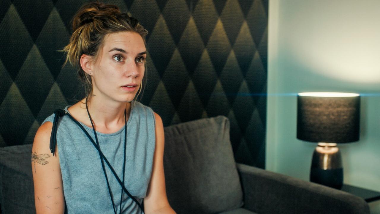 Maya Unger sitzt auf einem dunkelgrauen Sofa in einem Raum mit dunkel gemusterter Wand und einer Tischlampe.