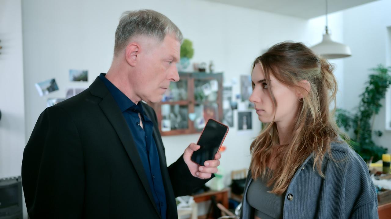 © ORF/MR Film/Christopher Glanzl Andreas Lust in dunklem Anzug zeigt Maya Unger ein Smartphone, beide stehen in einem wohnlich eingerichteten Raum.