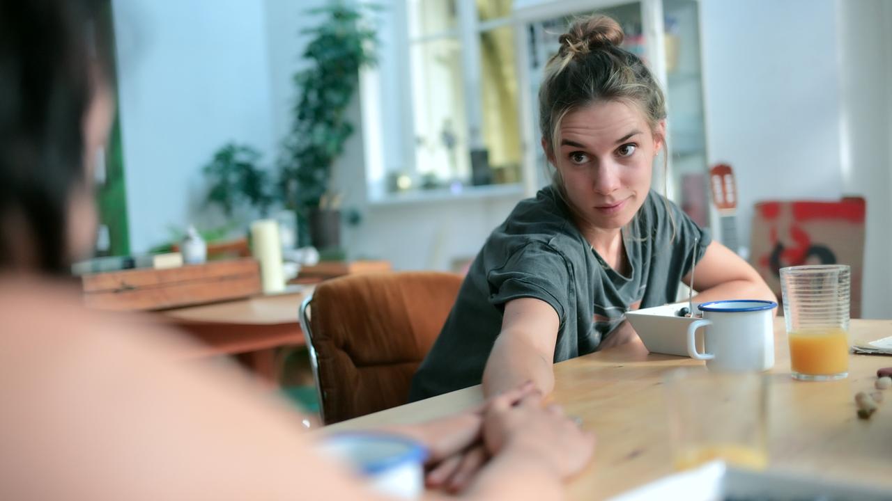 © ORF/MR Film/Christopher Glanzl Maya Unger sitzt mit einer weiteren Person an einem Holztisch und reicht dieser ihre Hand, neben ihnen stehen Getränke und Frühstücksschalen.