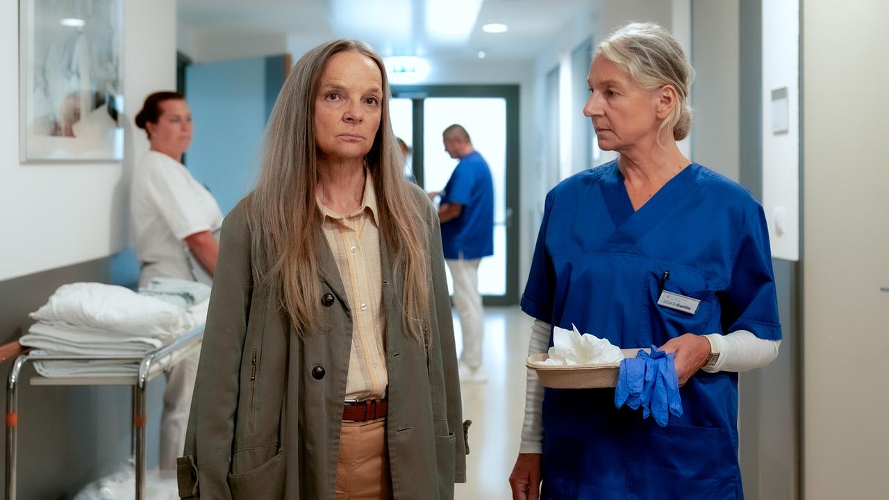 © ORF/MR Film/Petro Domenigg Sissy Höfferer im Mantel spricht im Krankenhausflur mit einer Krankenschwester in blauer Uniform, im Hintergrund arbeiten Pflegekräfte.