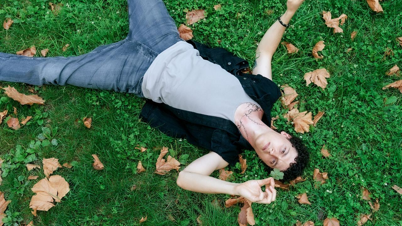 © ORF/MR Film/Petro Domenigg Etienne Halsdorf liegt mit dem Rücken ausgestreckt auf einer Wiese und hält ein vierblättriges Kleeblatt