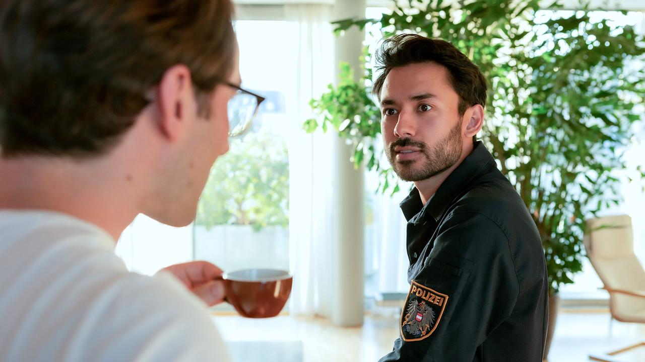 © ORF/MR Film/Anjeza Cikopano Tobias Resch mit einer Tasse Kaffee in der Hand blickt zu Harwin Kravitz in Polizeiuniform in einem lichtdurchfluteten Raum.