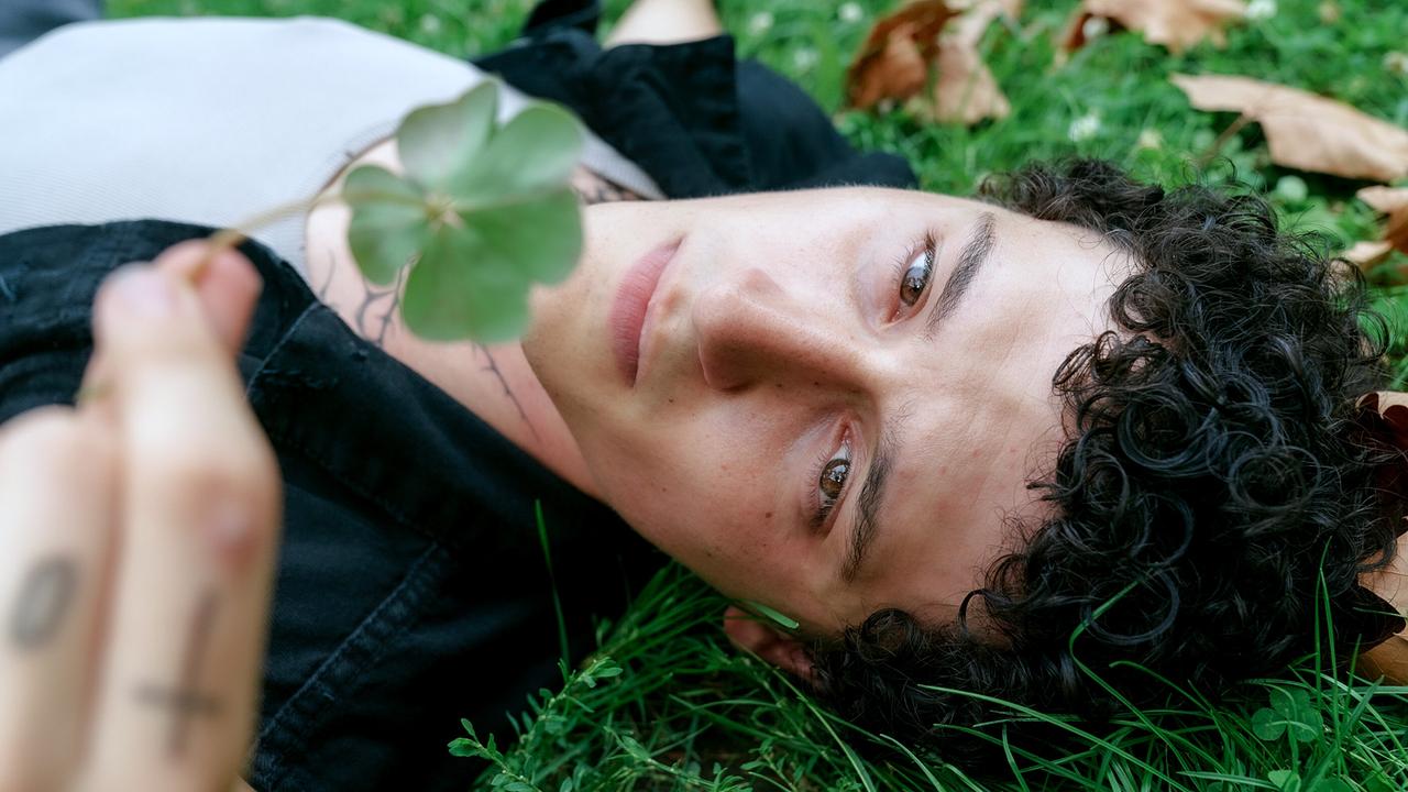 © ORF/MR Film/Petro Domenigg Etienne Halsdorf betrachtet auf einer Wiese liegend ein vierblättriges Kleeblatt, das er in seine Hand hält.