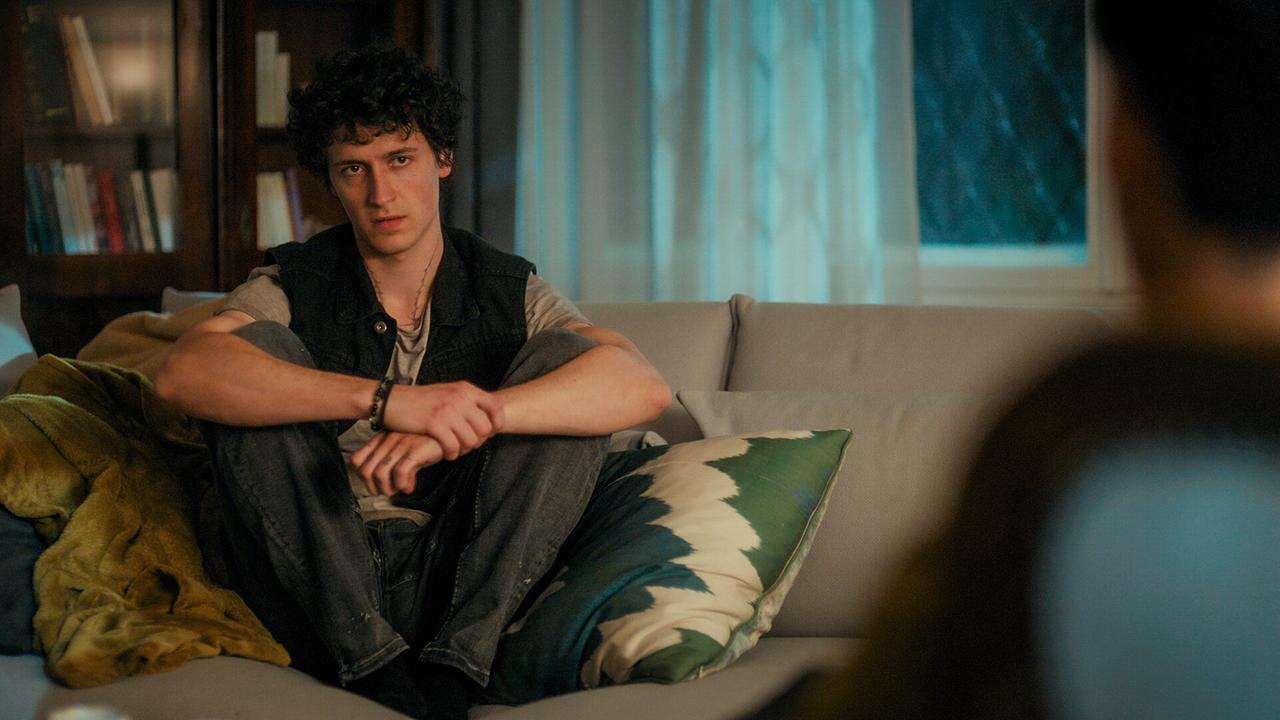 © ORF/MR Film Etienne Halsdorf sitzt abends auf einer Coach in einem Wohnzimmer; gegenüber jemand unscharf im Vordergrund.