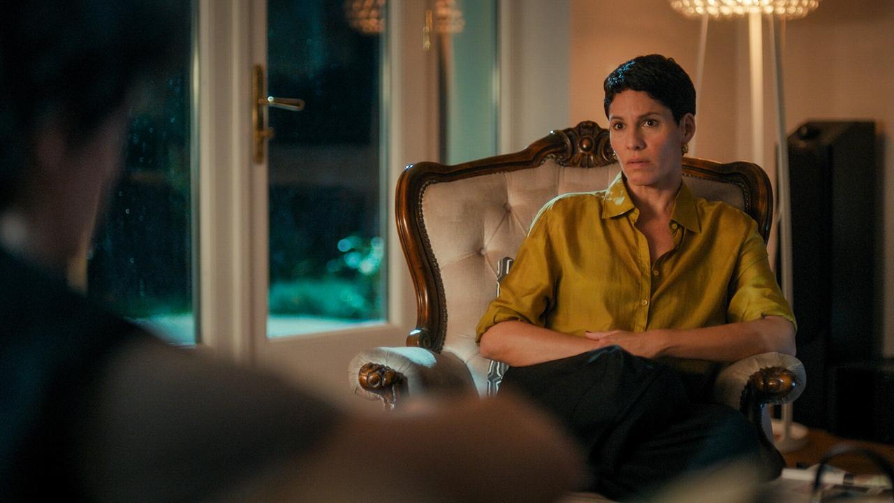 © ORF/MR Film Jasmin Gerat in gelbem Hemd sitzt abends in einem Sessel in einem Wohnzimmer, gegenüber jemand unscharf im Vordergrund.