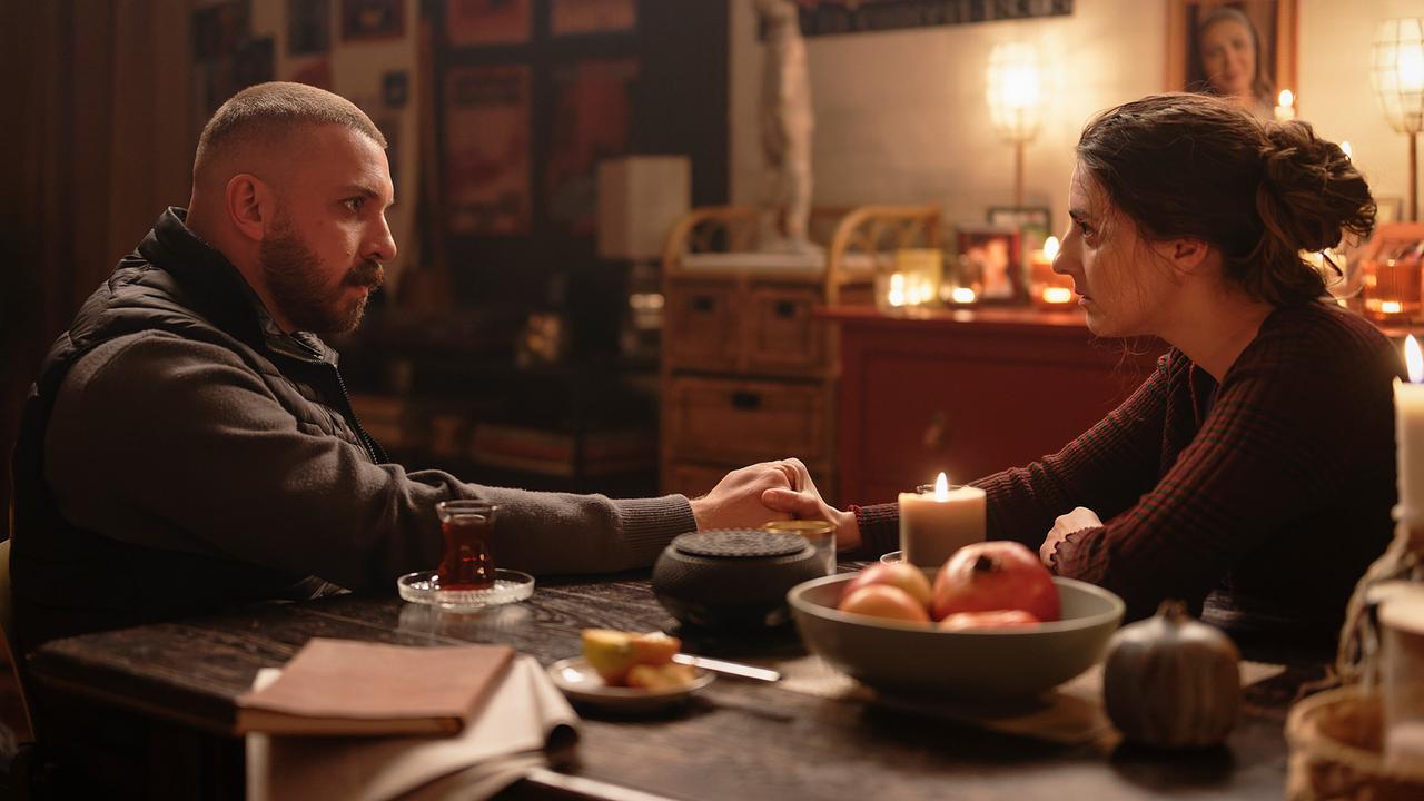 An einem gemütlich beleuchteten Tisch in einem Wohnraum sitzen sich Hamza Kulina und Almila Adak gegenüber und halten bei Kerzenlicht die Hände.