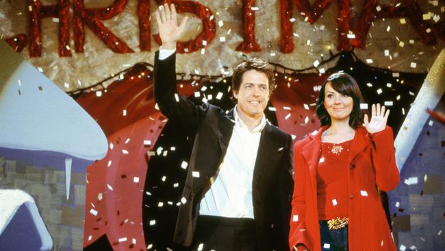 Ein Mann im schwarzen Anzug und eine Frau in rotem Mantel stehen unter herabfallendem Konfetti und winken, im Hintergrund weihnachtliche Dekoration mit rotem Schriftzug