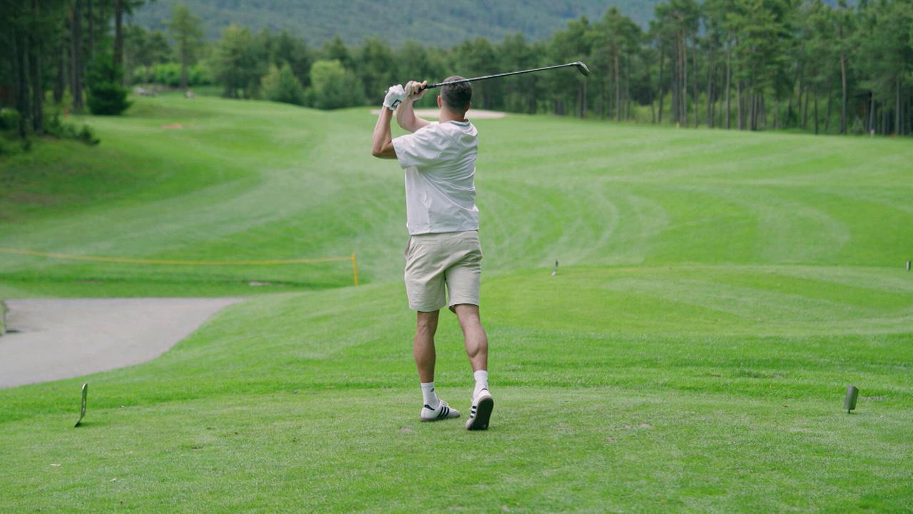 Ein Golfer schlägt einen Ball ab und hält den Schläger nach dem Schwung hoch, während sich der Fairway vor ihm öffnet.
