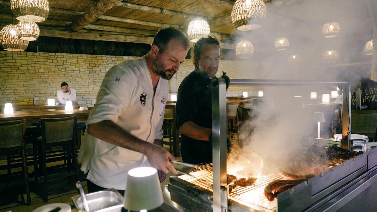 Zwei Köche bereiten in einem rustikalen Restaurant Speisen auf einem Grill mit offenen Flammen zu.