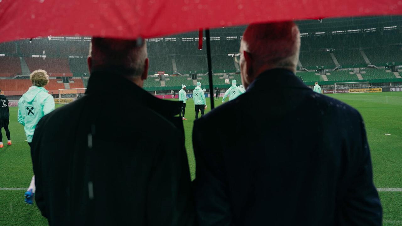 Bundespräsident Alexander Van der Bellen und ein weiterer Mann beobachten unter einem Regenschirm das Fußballtraining im Stadion.