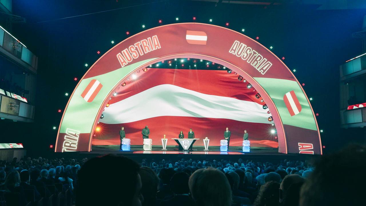 Eine große Bühne mit Publikum bei der WM-Auslosung in Washington zeigt das Podium unter einem Bogen mit der Aufschrift „Austria“ und einer rot‑weiß‑roten Flagge.