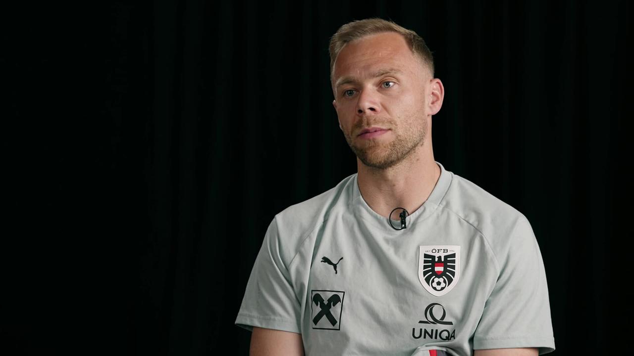 Alexander Schlager in Sportkleidung wird in einem Studio‑Setting vor schwarzem Hintergrund interviewt.