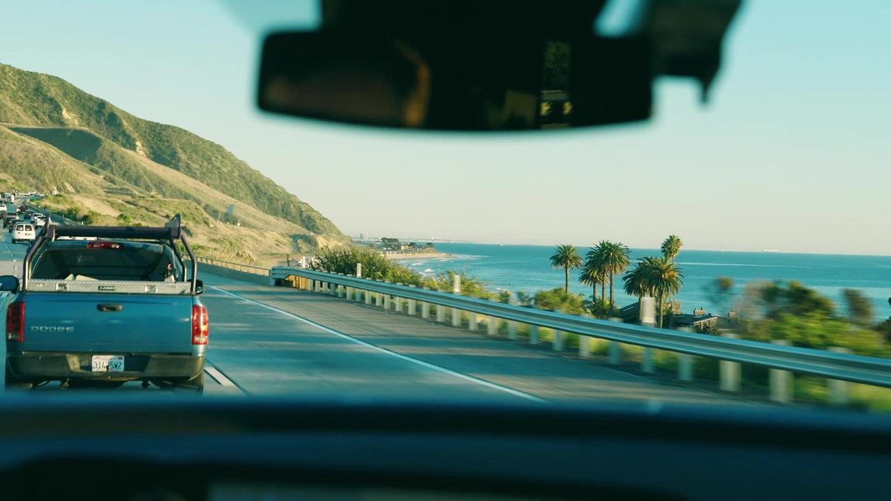 Eine Autofahrt entlang einer Küstenstraße an der Pazifik-Küste in Santa Barbara zeigt das Meer, Palmen und Hügel im Hintergrund.