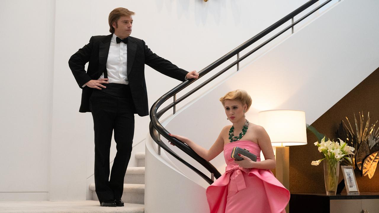 Donald Trump steht im Smoking auf einer Treppe und schaut zu Ivana Trump, die ihn in einem rosafarbenen Abendkleid bereits am Ende der Treppe erwartet