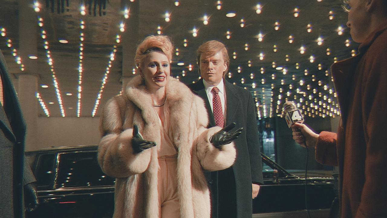 Maria Bakalova als Ivana Trump in einem hellen Pelmantel und Sebastian Stan als Donald Trump in einem dunkelgrauen Mantel haben ihre schwarze Limousine verlassen und werden von Reportern vor dem Eingang eines Gebäudes erwartet
