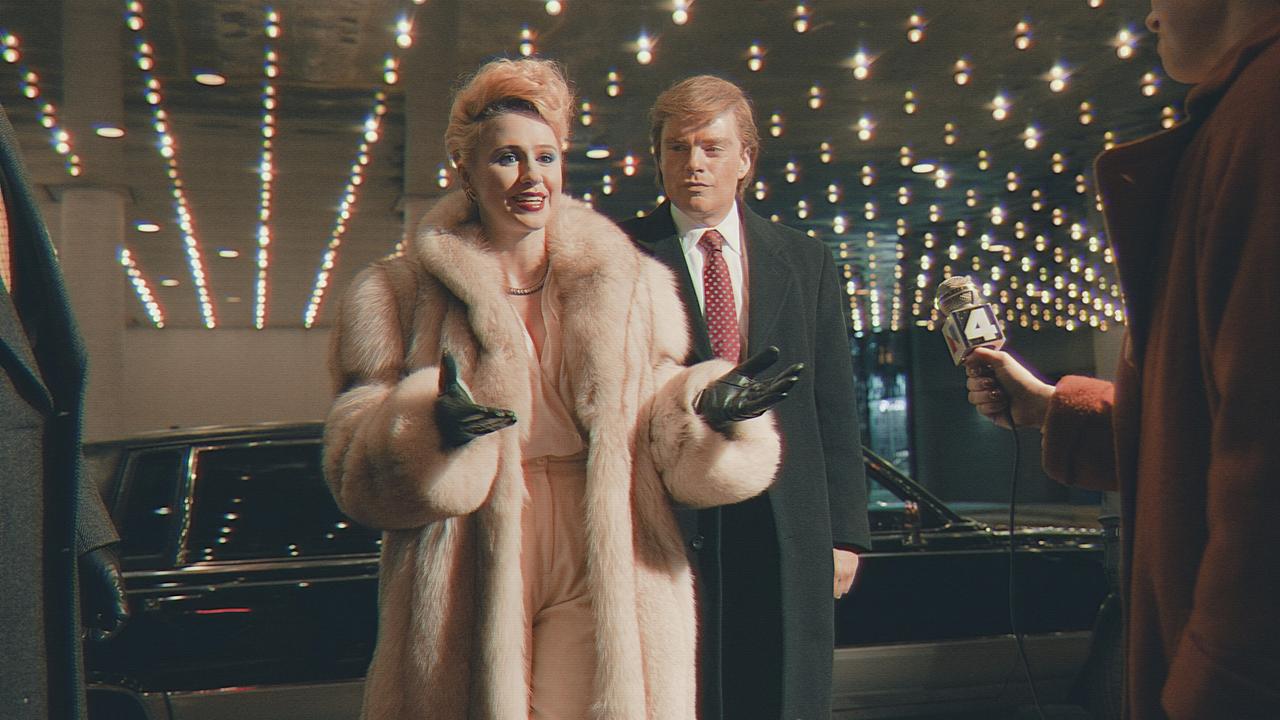 Maria Bakalova als Ivana Trump in einem hellen Pelmantel und Sebastian Stan als Donald Trump in einem dunkelgrauen Mantel haben ihre schwarze Limousine verlassen und werden von Reportern vor dem Eingang eines Gebäudes erwartet