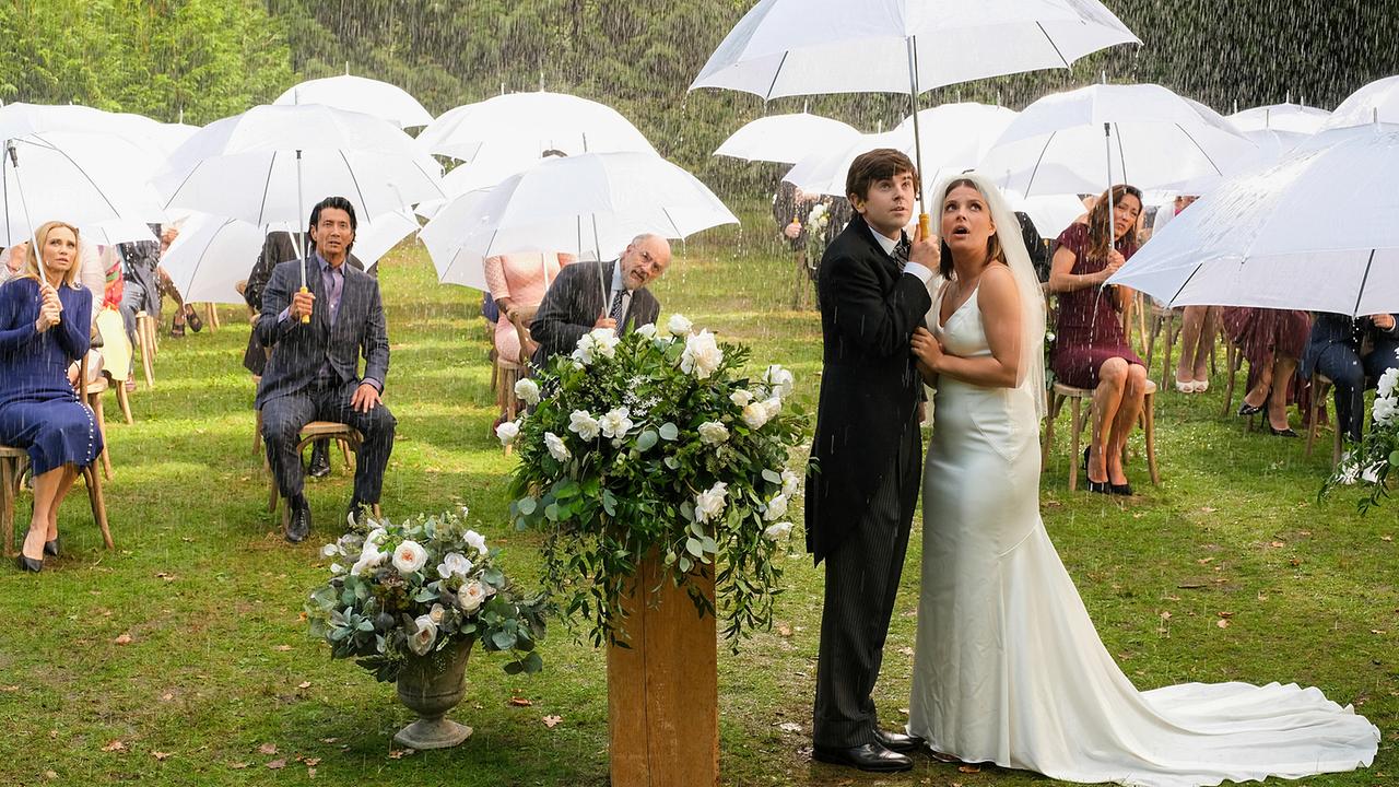 © ORF/Sony Pictures/ABC/Jeff Weddell Shaun und Lea stehen im Freien in einem starken Regen in feierlicher Bekleidung, hinter ihnen sitzen Kollegen und Kolleginnen auf Stühlen mit Abstand, das Paar und alle anderen halten große durchsichtige Regenschirme in der Hand
