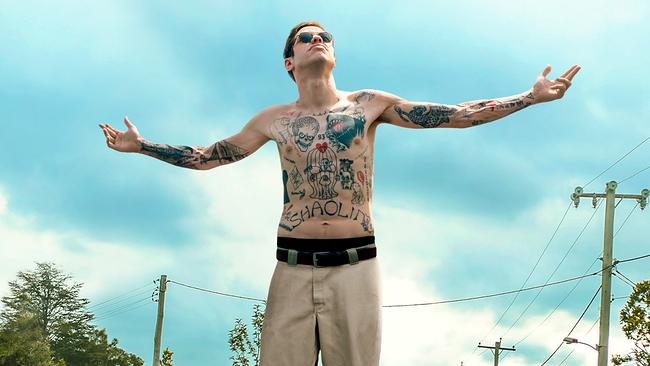 Ein junger Mann auf einer Straße, mit nacktem, tätowiertem Oberkörper breitet er seine Arme aus und blickt mit dunkler Sonnenbrille hoch in den blauen Himmel