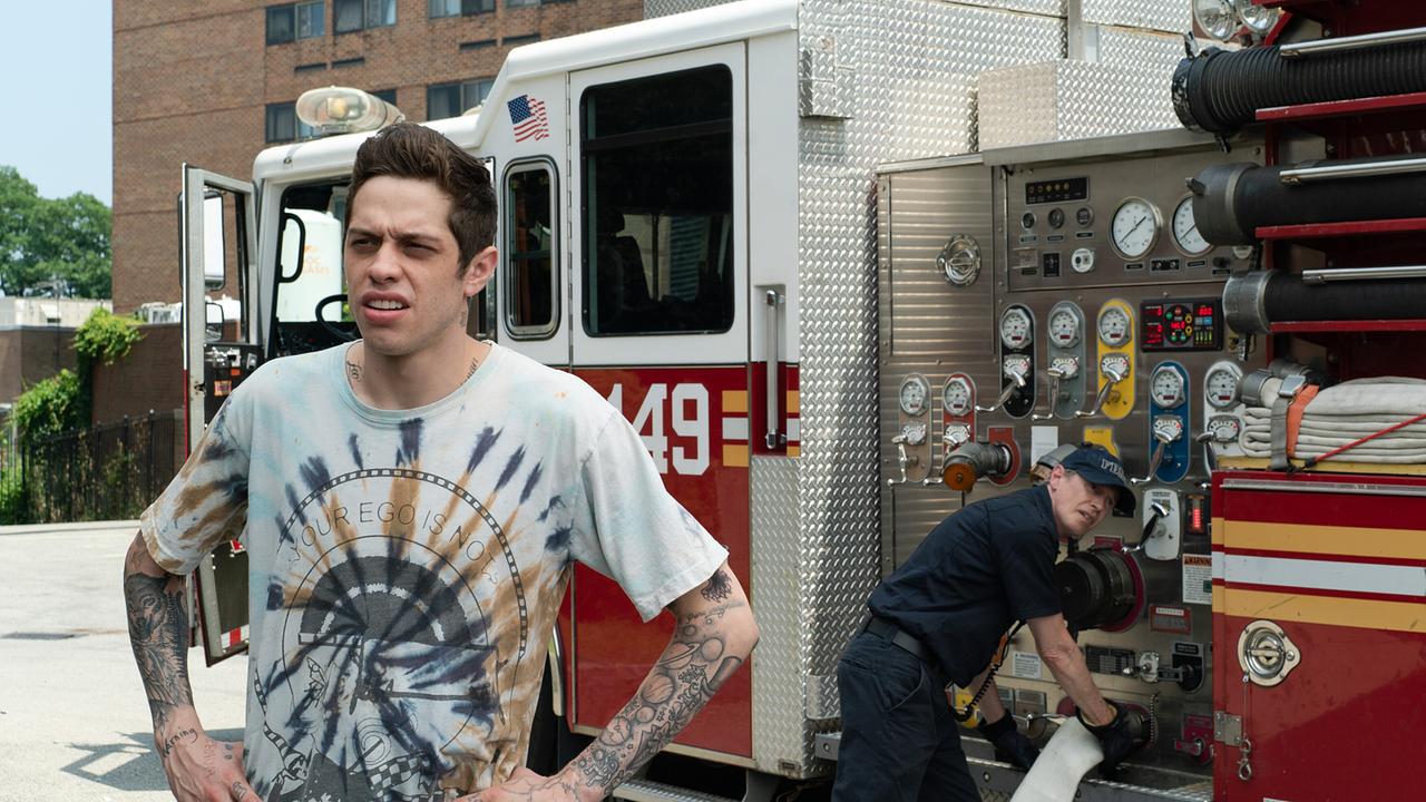 Ein junger Mann in einem hellen, gebatikten T-Shirt steht mit den Händen an seiner Hüfte vor einem stehenden Feuerwehrwagen, ein Feuerwehrmann montiert gerade den Schlauch am Wagen