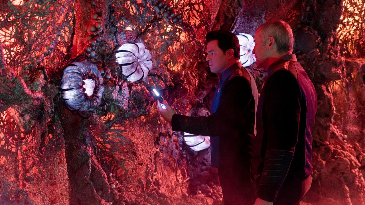 Zwei Männer in futuristischen Uniformen untersuchen mit leuchtenden Geräten eine rot beleuchtete, organisch wirkende Wandstruktur mit weißen, blütenartigen Formen