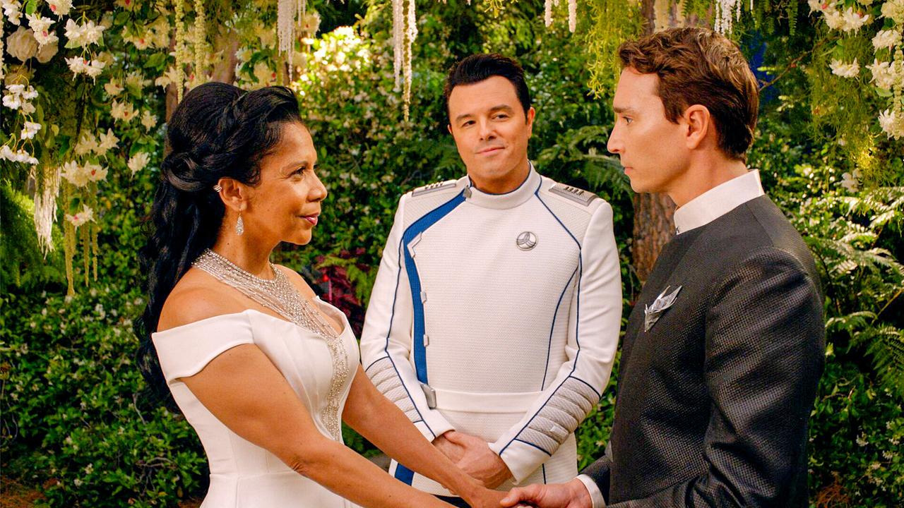 © ORF/ProSieben/20th Century Studios/Hulu/Gilles Mingasson Eine Frau im weißen Kleid hält die Hände eines Mannes im grauen Anzug, hinter dem Paar steht ein lächelnder Mann in festlicher, weißer Uniform, im Hintergrund Pflanzen und blühende Blumen