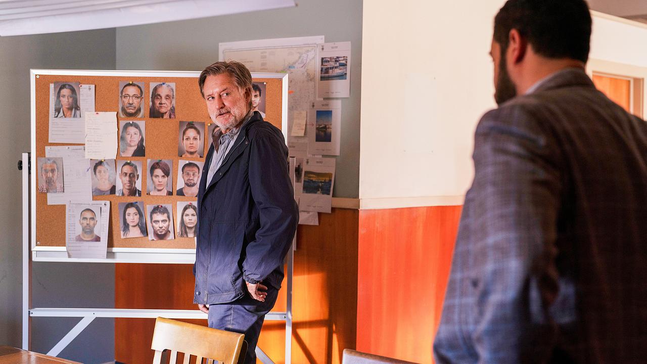 Harry Ambrose steht vor einer Tafel, auf der die Porträtfotos der in den Fall involvierten Personen gepinnt sind, und spricht mit Detective Brookes