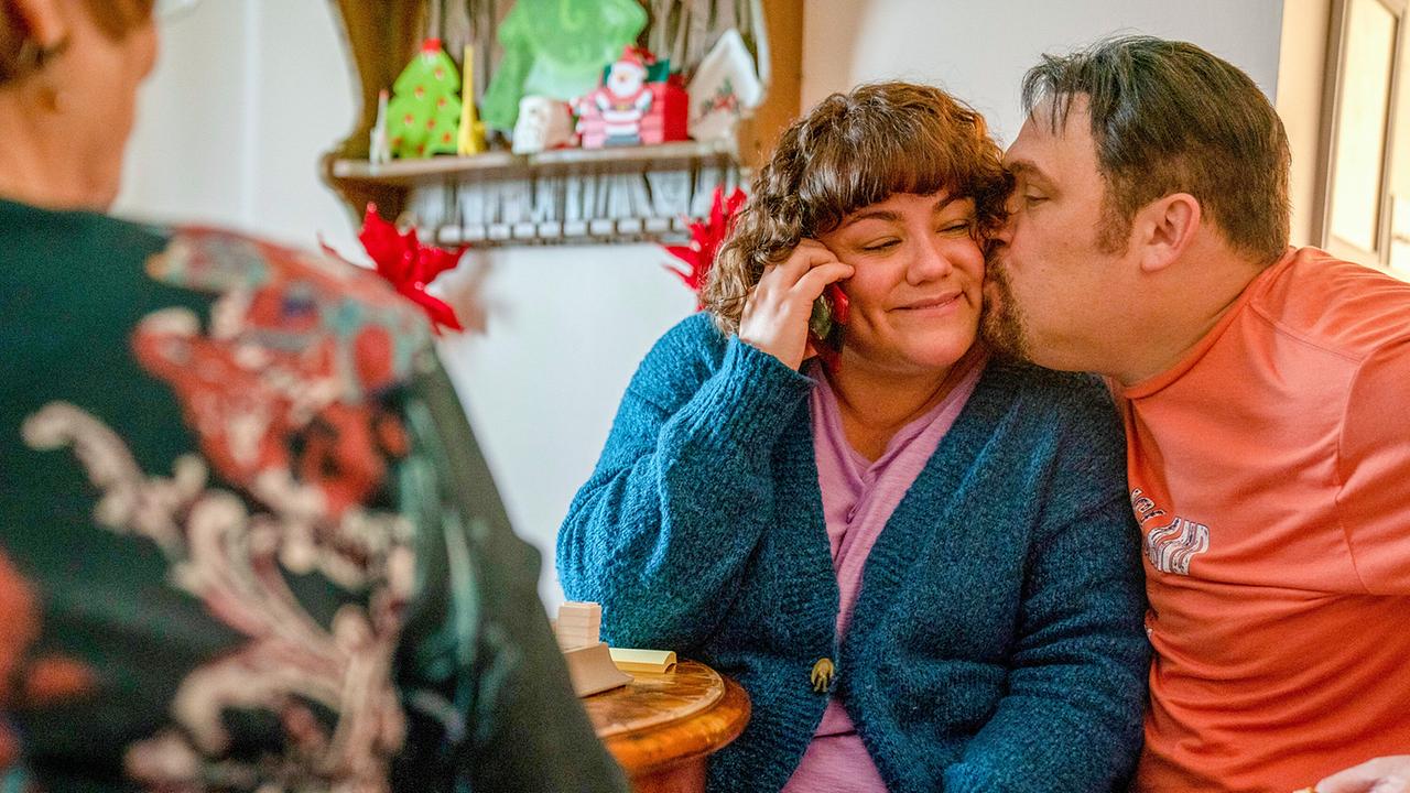 © ORF/Universal/NBC/Skip Bolen Betsy Faria in lilafarbenem Shirt und blaugrüner Strickjacke sitzt glücklich lächelnd neben Russ Faria, der ihr einen Kuss auf die Wange drückt