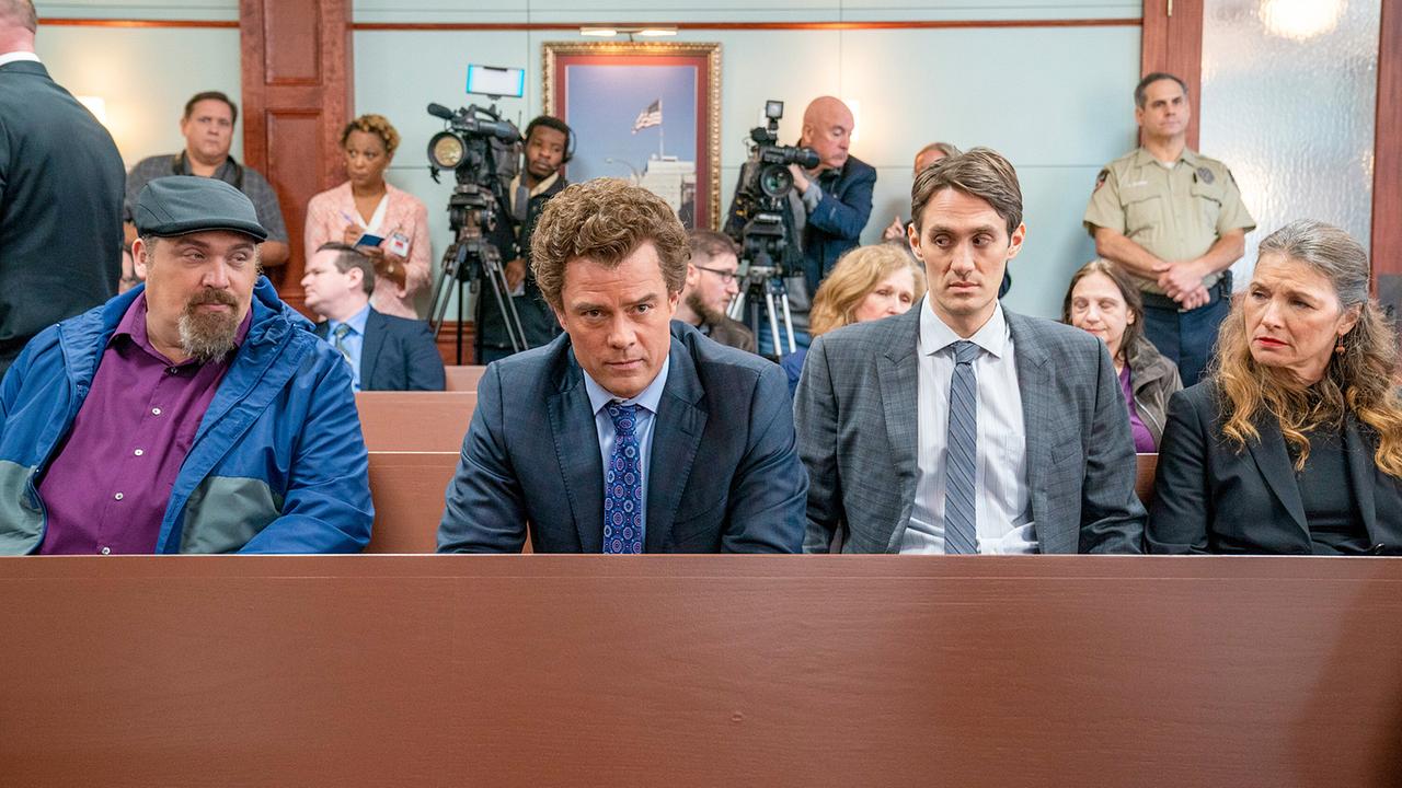 Drei Männer und eine Frau sitzen in der ersten Reihe bei einem Gerichtsprozess, hinter ihnen stehen Kameramänner mit Kameras an der Wand