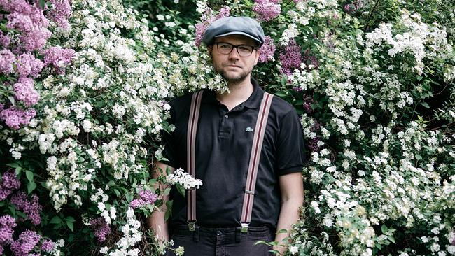 Thomas Stipsits mit Kappe und Hosenträgern steht vor dichtem, blühendem Fliederstrauchwerk mit weißen und violetten Blüten.