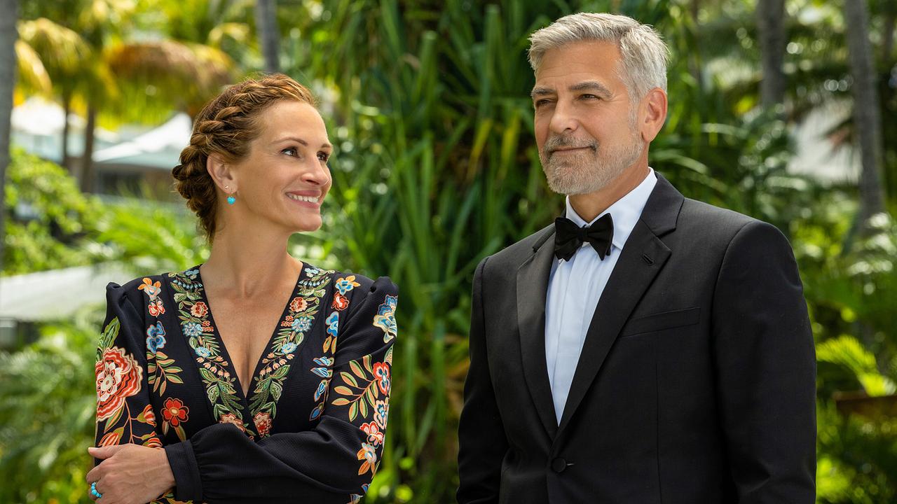 Julia Roberts als Georgia Cotton in einem schwarzen Kleid mit bunten floralen Apllikationen und George Clooney als David Cotton im eleganten Smoking, freundlich lächelnd stehen die beiden nebeneinander
