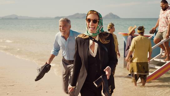 Julia Roberts als Georgia Cotton in schwarzem Hosenanzug und türkisem Kopftuch und George Clooney als David Cotton in beiger Hose und blauem Hemd mit den Schuhen in der Hand auf einem paradiesischen Sandstrand verlassen sie lachend ein gelandetes Boot