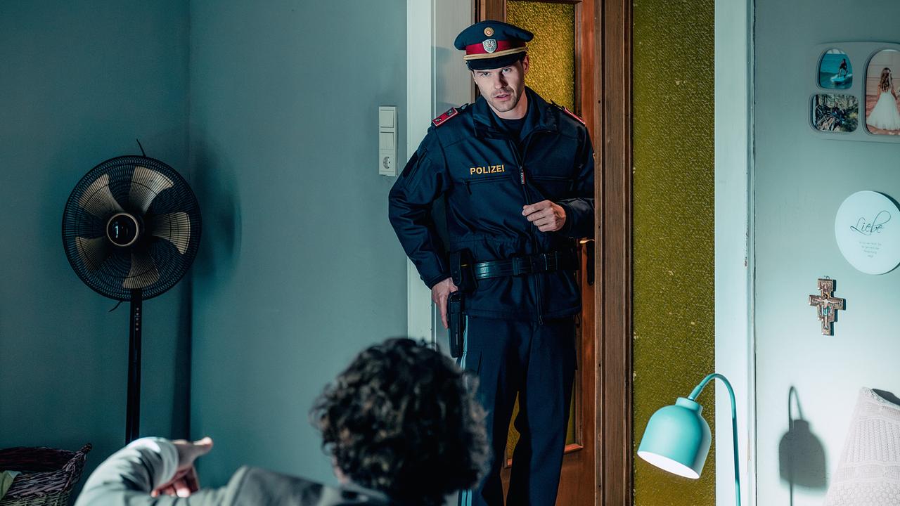 © ORF/Rundfilm/Christoph Karl Izmenyi Tobi Gruber betritt in Polizeiuniform ein Wohnzimmer, mit der rechten Hand hält er die Pistole im Halfter, er spricht mit einem Mann, der ihm gegenüber auf einer Couch sitzt