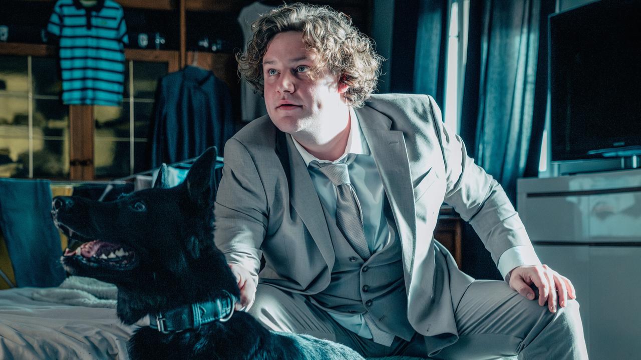 © ORF/Rundfilm/Christoph Karl Izmenyi Ein Mann sitzt im hellgrauen Anzug mit Weste in einem Zimmer auf einem Bett und hält einen Hund an dessen Halsband, er und der Hund blicken in dieselbe Richtung