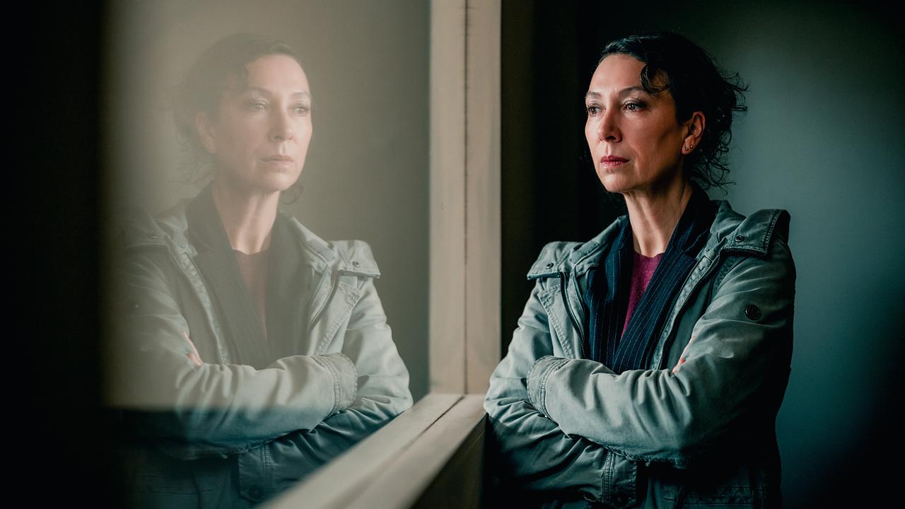 © ORF/Rundfilm/Nikolett Kustos Ursula Srauss steht als Fanny Gruber mit verschränkten Armen in einem abgedunkelten Raum vor einem Fenster, in der sie sich spiegelt, ernst und gefasst blickt sie durch die Scheibe ins Verhörzimmer