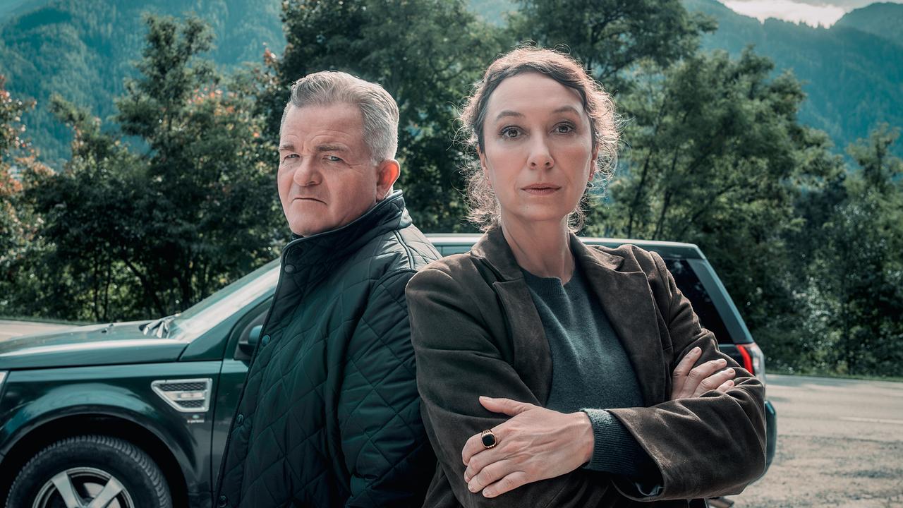 © ORF/Rundfilm/Nikolett Kustos Ursula Strauss als Fanny Gruber und Cornelius Obonya als Benedikt Renner stehen vor einem grünen Geländewagen und blicken direkt in die Kamera, im Hintergrund Bäume und bewaldete Berge
