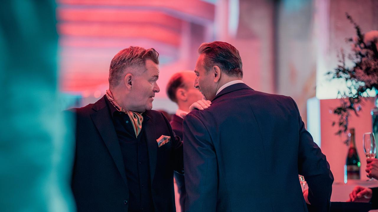 © ORF/Rundfilm/Nikolett Kustos Cornelius Obonya als Benedikt Renner bei einer Abendveranstaltung im Gespräch mit einem Geschäftsmann im Anzug und hält seine Hand auf dessen Schulter