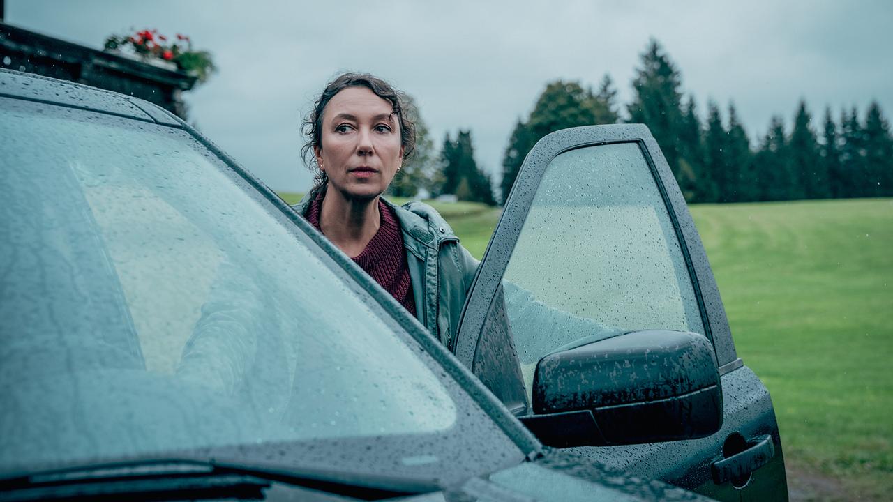 © ORF/Rundfilm/Nikolett Kustos Fanny Gruber steht bei Regen vor der geöffneten Autotür und blickt über eine ländliche Wiese.