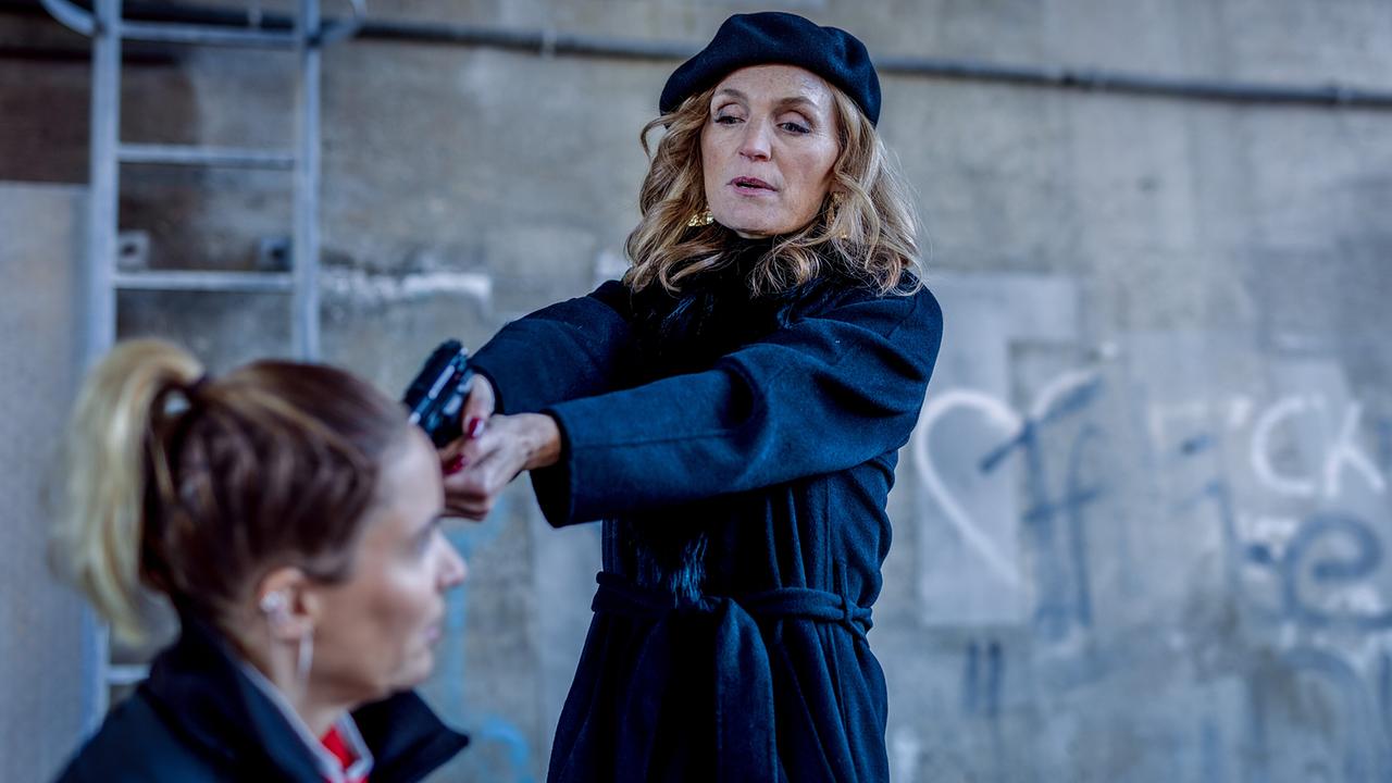 Caterina Relli zielt mit einer Pistole auf Lisa Kuen unter einer Brücke vor graffitibeschmierten Wänden.