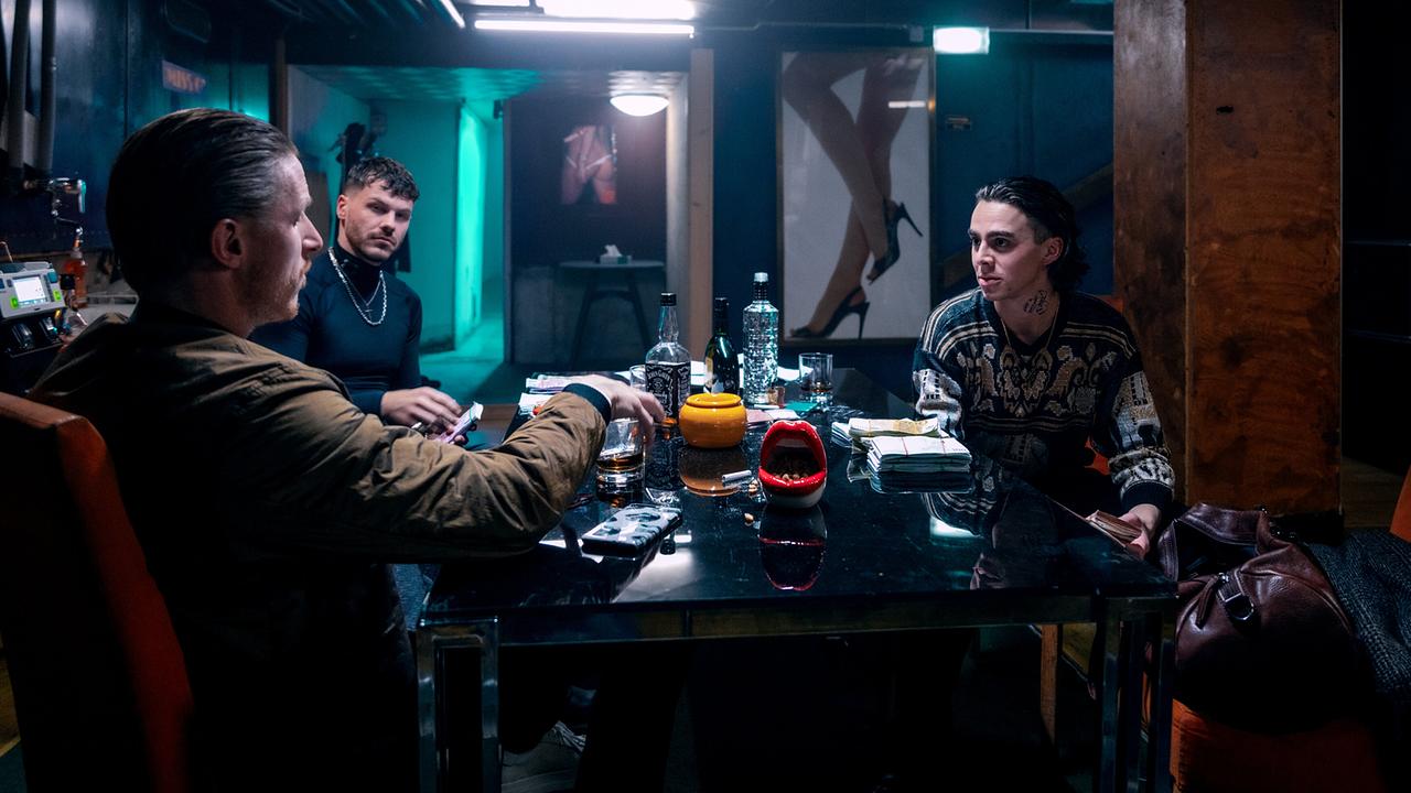 Albert Mosel, Luan Kuka und Marco Relli sitzen an einem dunklen Tisch, auf dem Geldbündel liegen und Alkoholflaschen stehen, in einem dunklen Raum.