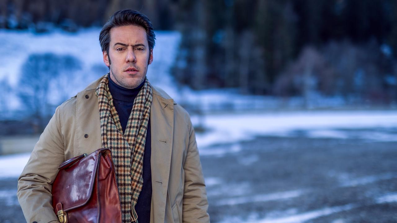 Alex Yüsüf-Demir in Mantel und Schal hält eine braune Aktentasche im winterlichen Freien.