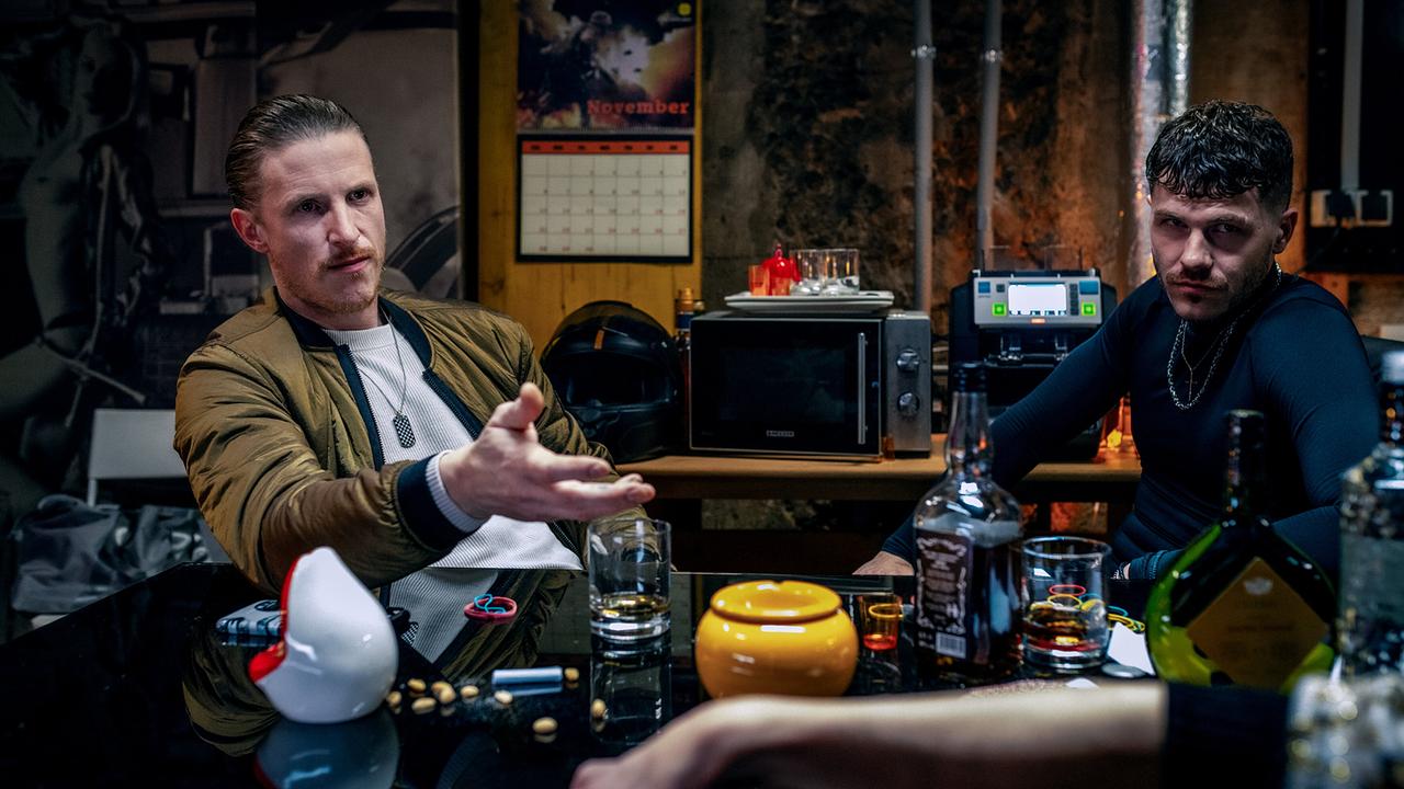 Albert Mosel und Luan Kuka sitzen an einem Tisch in einem Kellerraum voller Alkoholflaschen und Geldscheine.