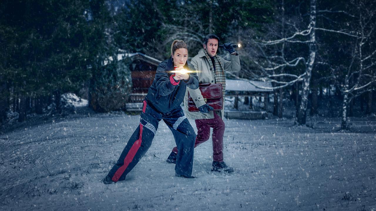 Lisa Kuen und Alex Yüsüf-Demir stehen im Schneegestöber auf einer Waldlichtung und leuchten mit Taschenlampen in Richtung eines Hauses.