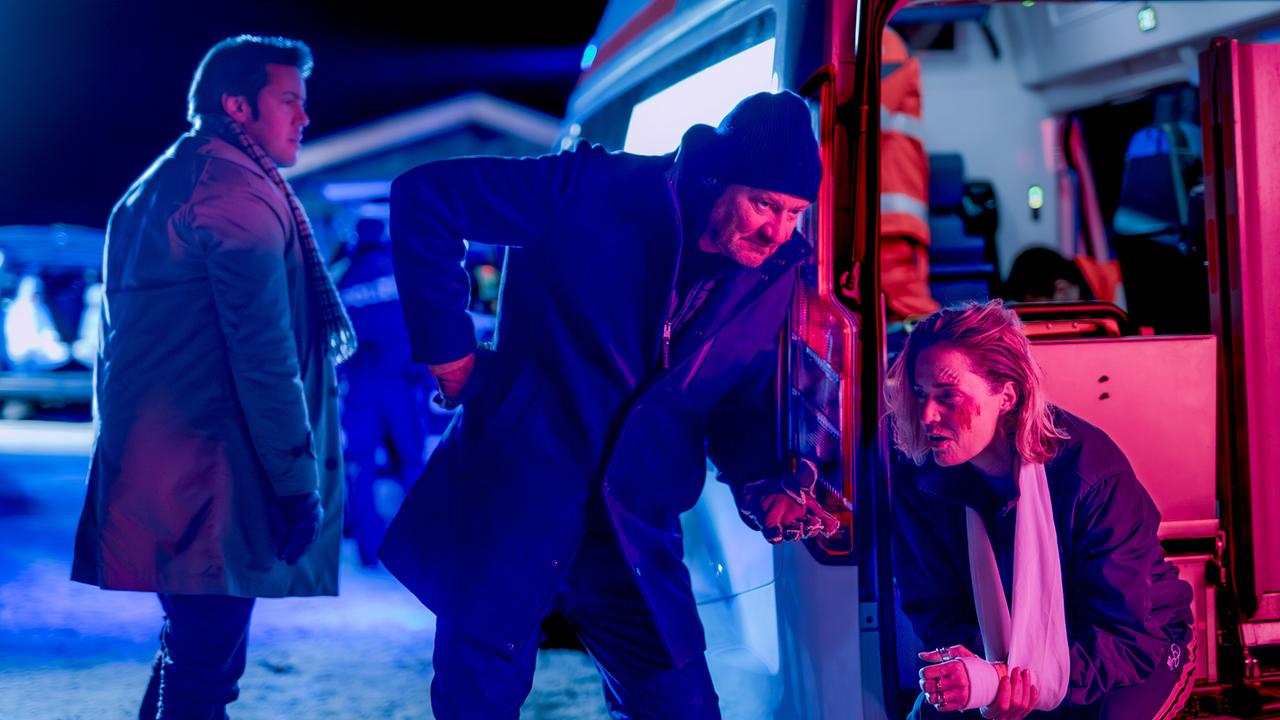 Alex Yüsüf-Demir und Oberst Günther Schupp stehen nachts vor einem Rettungswagen, während Lisa Kuen verletzt und erschöpft sich an den Rettungswagen stützt.